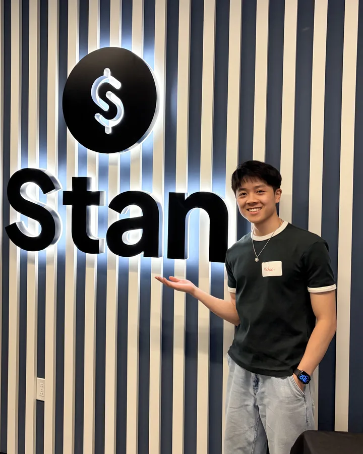 Michael smiling and gesturing at a lit Stan sign on a striped wall.